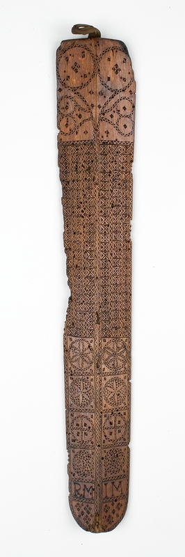 A long, slender wooden artefact with intricate patterns carved into it, which are especially densely carved in the upper centre.