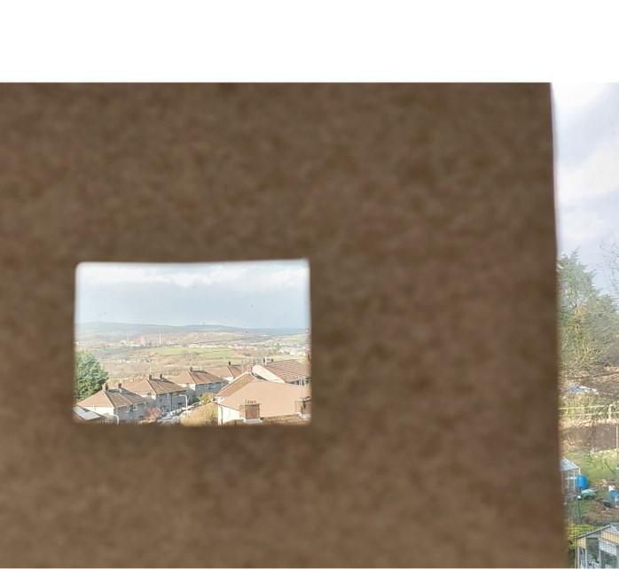 Photo of someone holding a cardboard viewfinder with houses and trees visible through small the rectangle gap