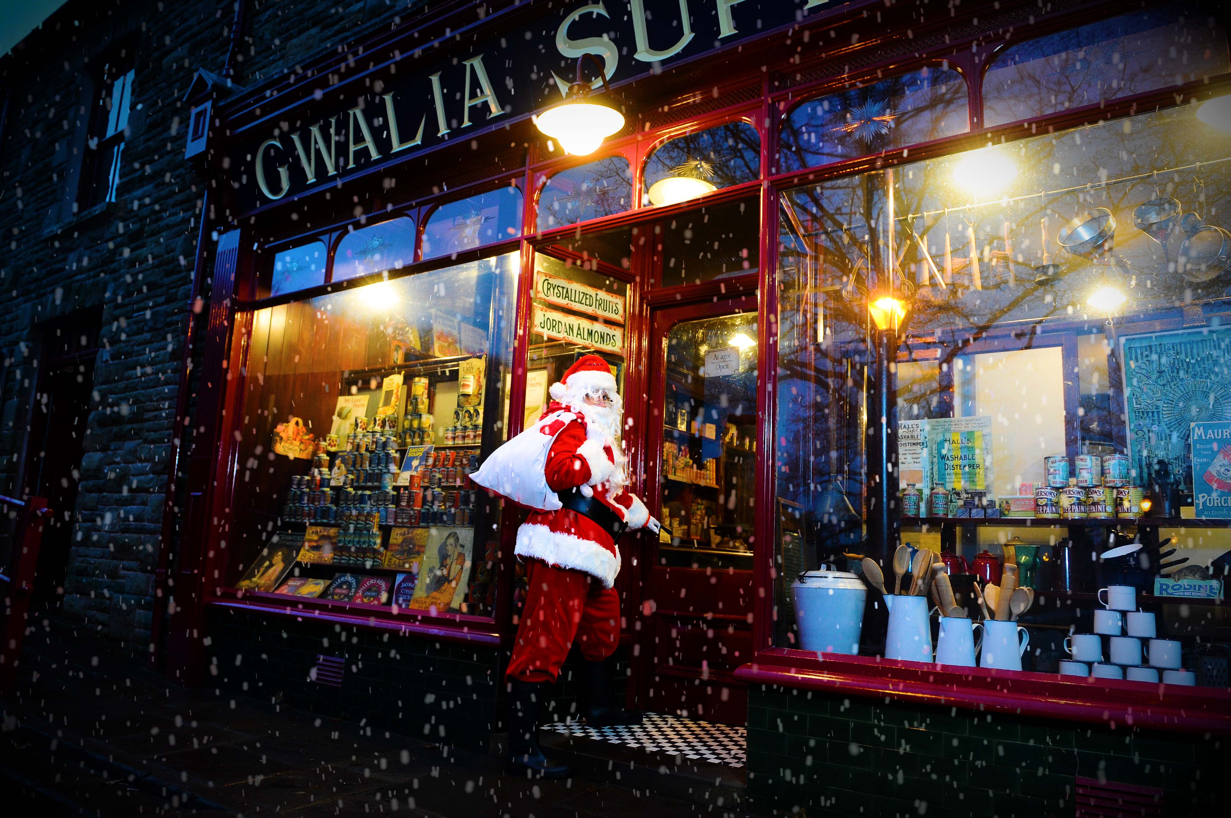 Father Christmas stands in a shop doorway while snow falls.