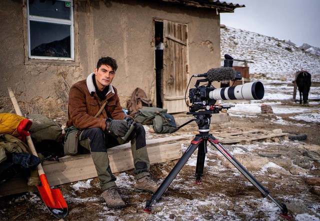 Man sat outside rustic building with filming equipment, light snow underfoot
