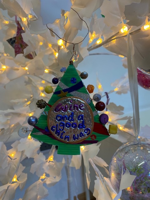 Photograph of a Christmas Tree decoration with a gold button in the middle with the words 'Wine and a good chin wag' written on it in purple and blue.