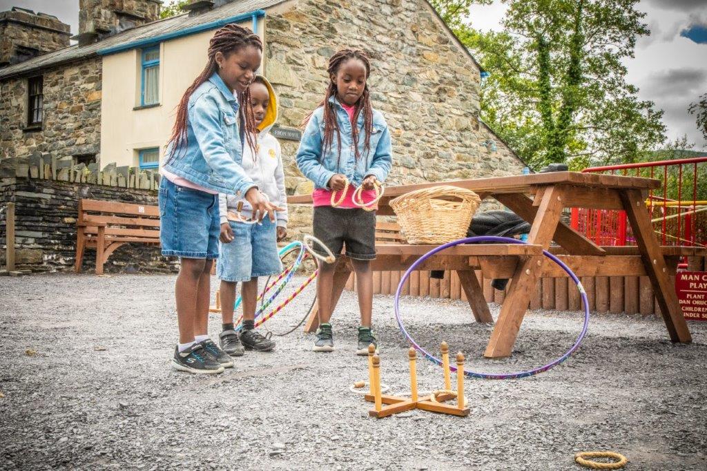 Three young children in denim playing an old game by throwing a circular rope at wooden pegs; there are hula hoops around them and a picnic bench