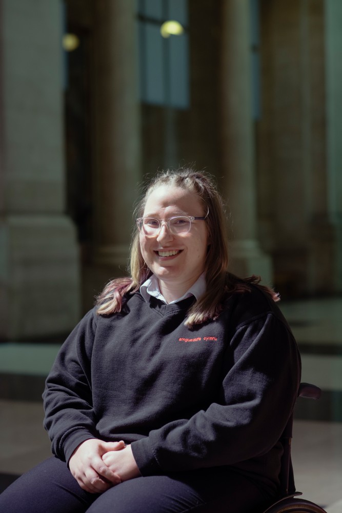 A young woman sat in a wheelchair smiling at the camera; she's wearing a navy Amgueddfa Cymru jumper and is sat in the main hall at National Museum Cardiff.