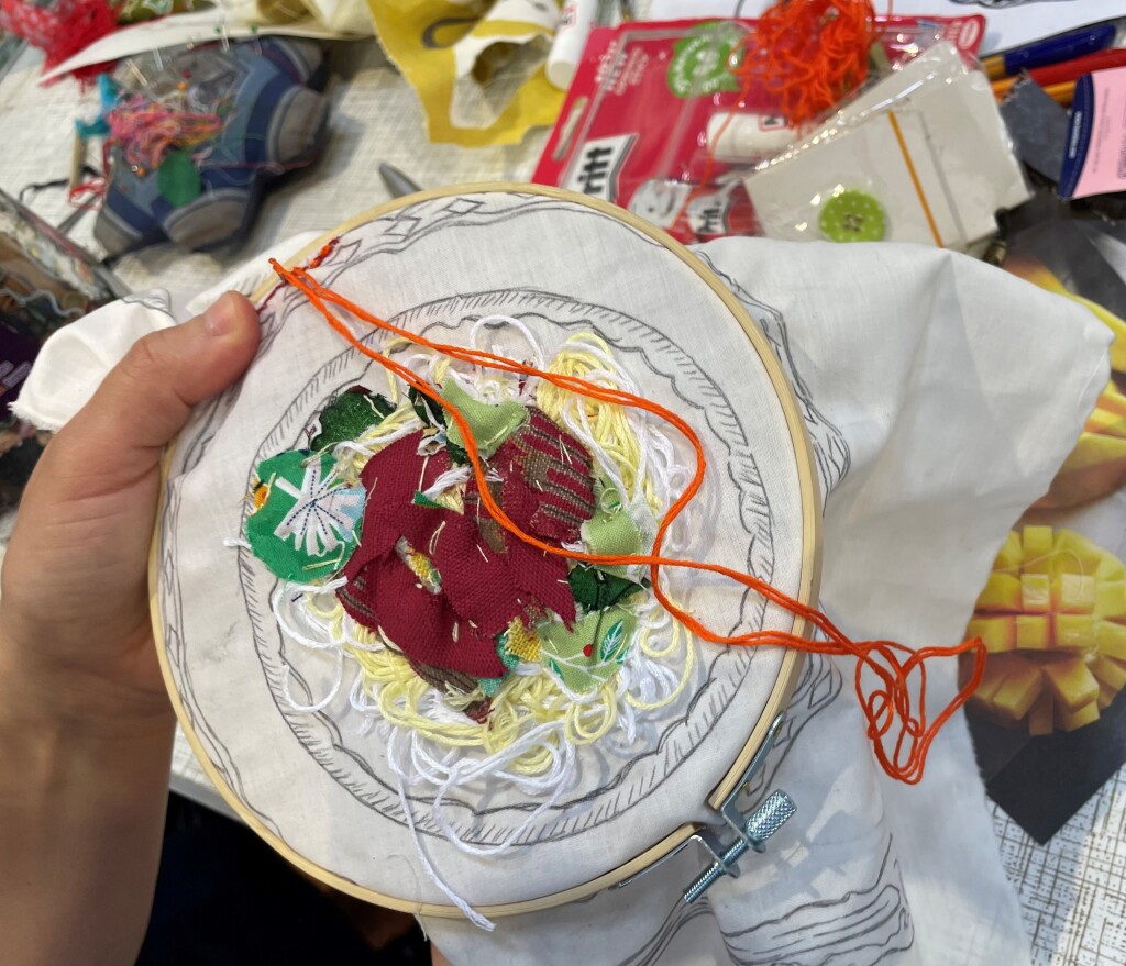 An embroidery hoop with yarn and textiles hand-stitched to depict a plate of spaghetti and tomato sauce