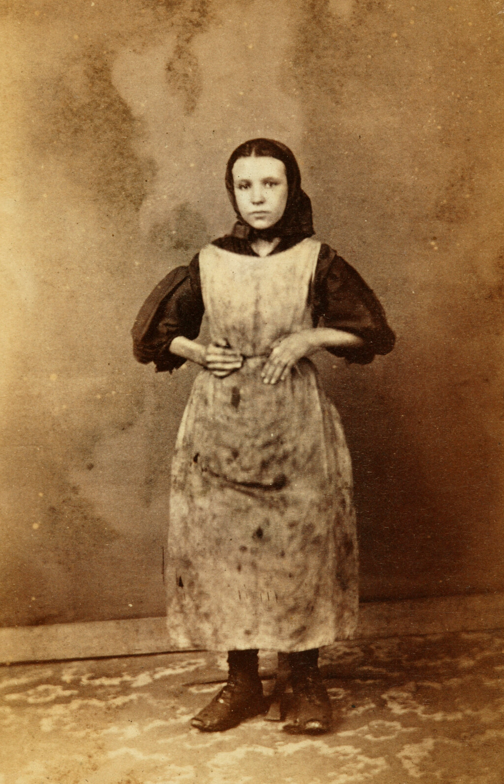 Old picture of a young woman standing with hands on hips while wearing a dirty apron and flared shirt underneath. 