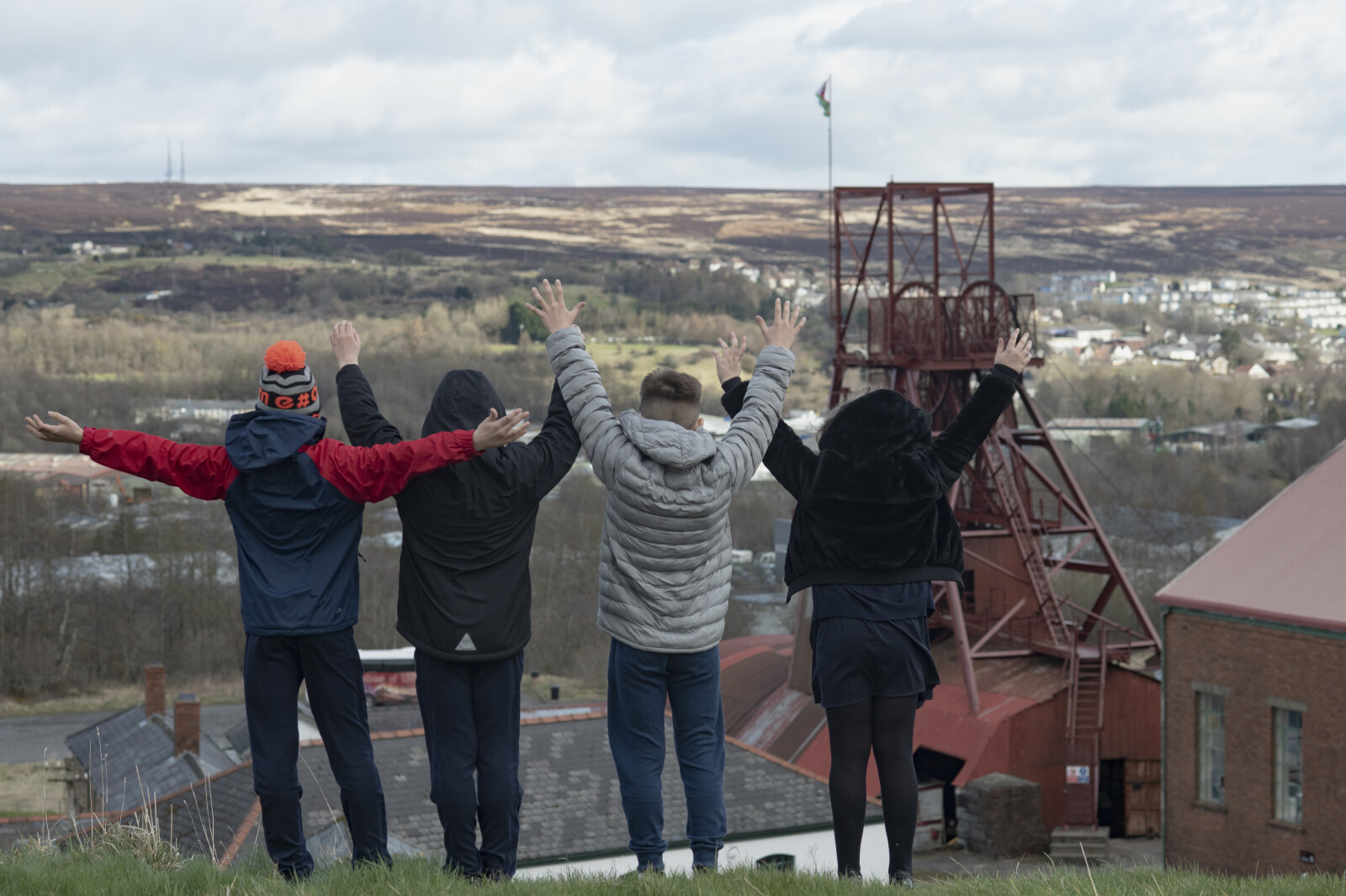Four young people standing with their backs to the camera and their hands up in the air as they look over the Big Pit Winding Wheel