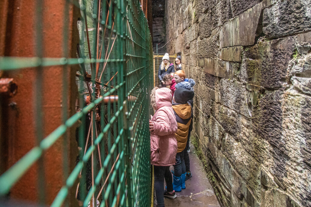 A group of children stand looking at a large metal waterwheel