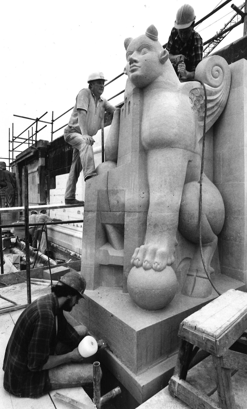 Black-and-white photograph of three workmen in hard hats working on a large stone sculpture of a winged mythical creature on the top storey of a building