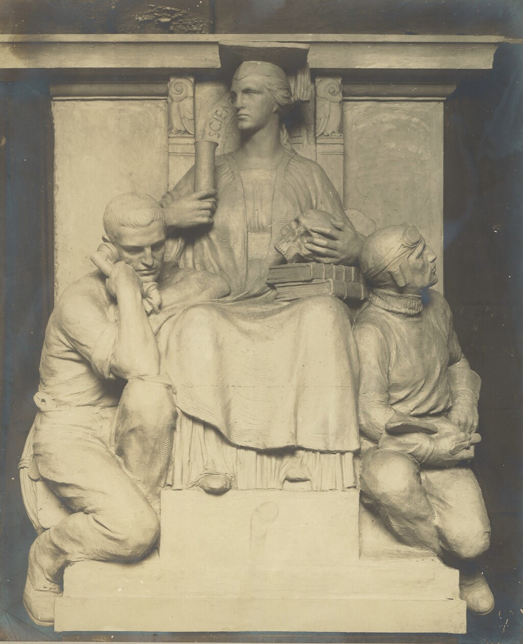 Sepia photograph of a plaster model for a sculpture of three figures: a seated woman wearing a square academic cap (mortar board) raised above two seated men either side of her, one holding a telephone receiver and the other dressed as a pilot