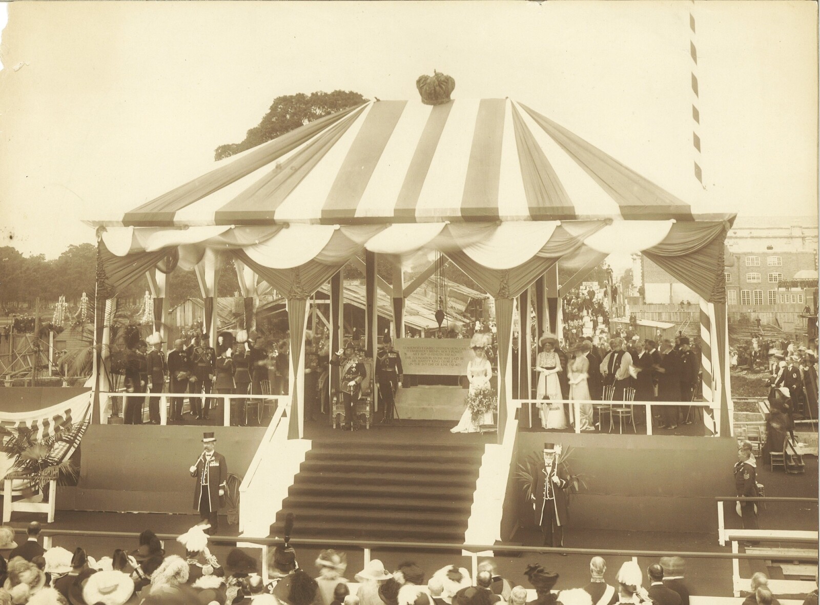 Sepia-toned photograph of King George V and Queen Mary standing under a striped canopy at the ceremony to lay the Foundation Stone. The ladies are in long pale dresses with large hats and the men are in ceremonial military dress