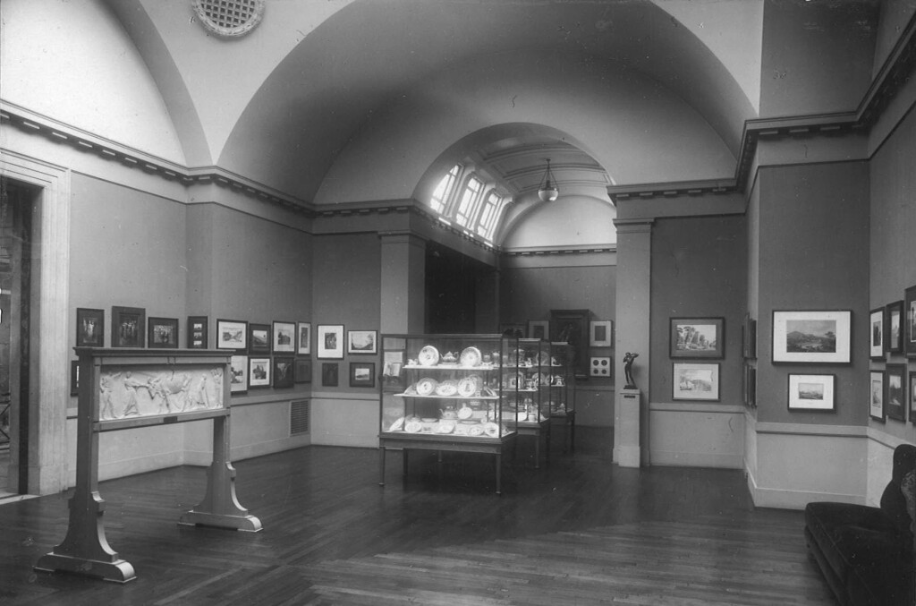 Photograph of the interior of the Pyke Thompson Gallery, framed pictures line the walls and a glass cabinet of Welsh China stands in the centre of the room