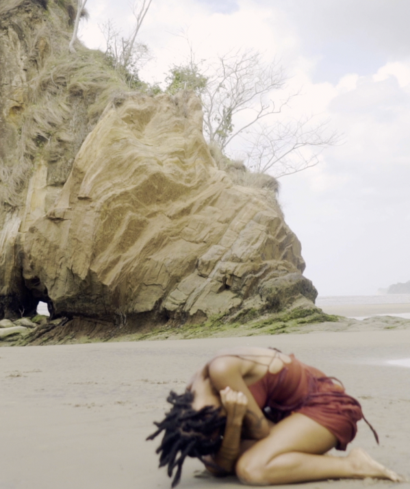 A still from Gesiye's film, showing a woman kneeling down on a beach with a rock formation behind her