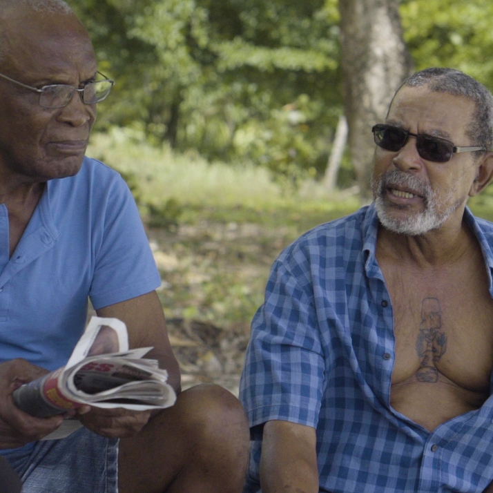 A still from Gesiye's film, showing two men in conversation. One wears a blue tshirt, the other a blue shirt with the buttons undone.