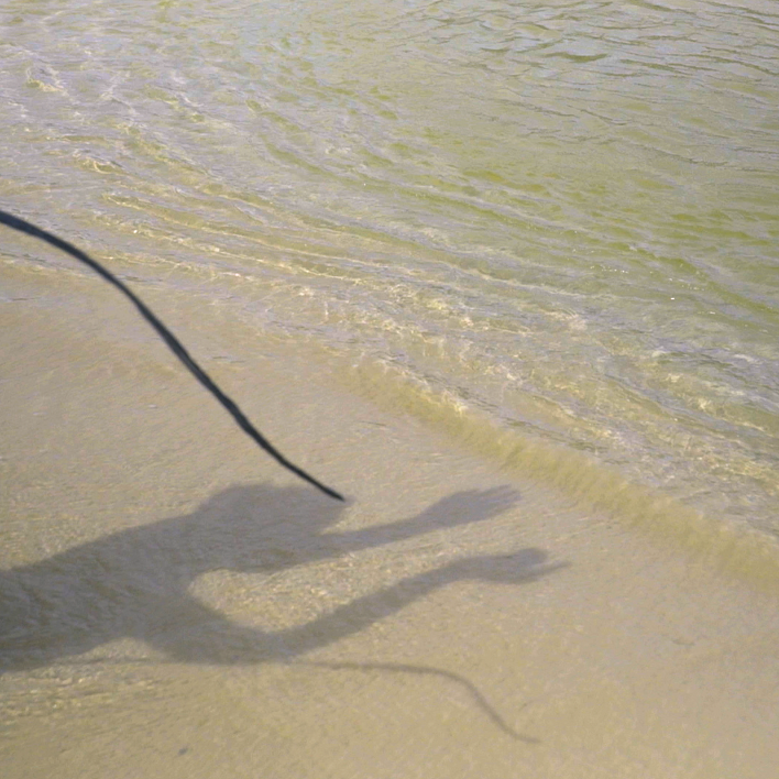 A still from Gesiye's film, showing a sandy beach, clear sea water and the shadow of a person dancing