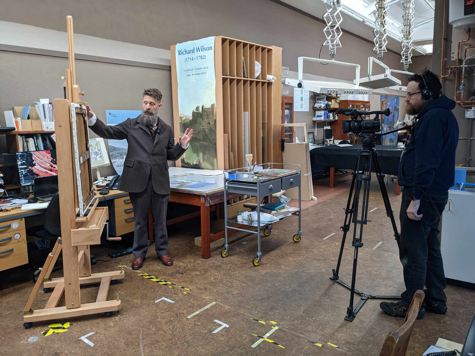 A man films another man standing in front of an artwork in an art conservation studio.