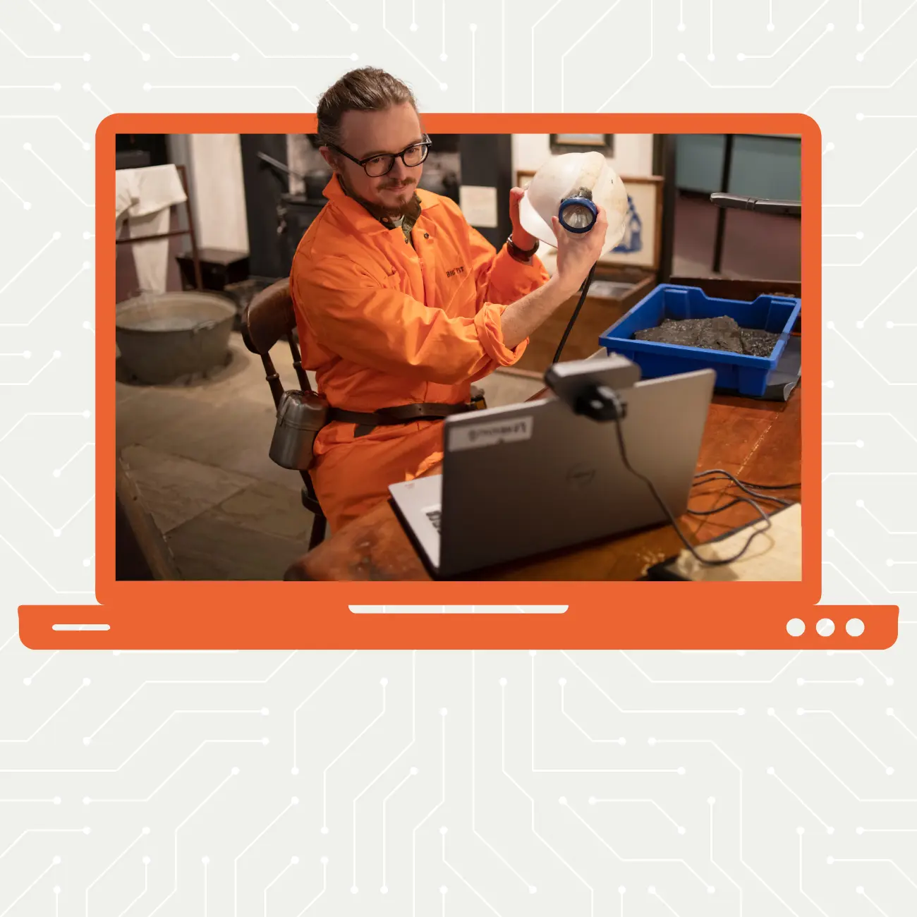 Man in an orange jumpsuit holding a helmet with a head torch up to a camera