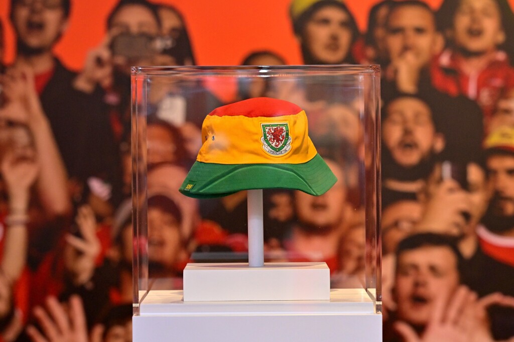 A Wales bucket hat in a Perspex case. The hat has three stripes - green, yellow and red. The Cymru badge is in the center of the yellow band. 