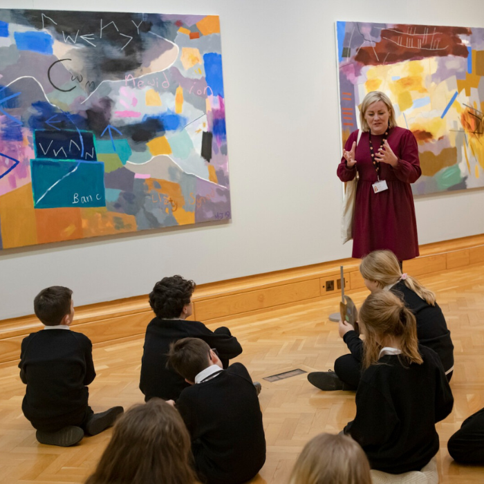 Photo of a learning facilitator speaking to a group of children in an art gallery. On the wall are two large colourful abstract paintings.