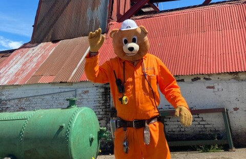 Butty Bear is standing in front of the red mining Wheels at Big Pit. He is giving the camera a thumbs up