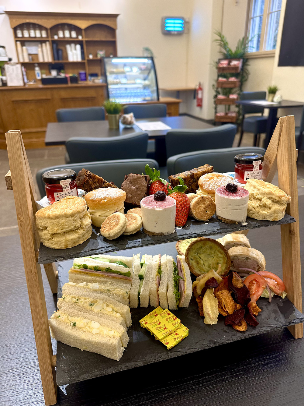 A slate shelf sits in the middle of a cafe table, on it sits a variety of cakes, baked goods and sandwiches including fluffy scones, smooth looking cheescake bites and sausage rolls. 
