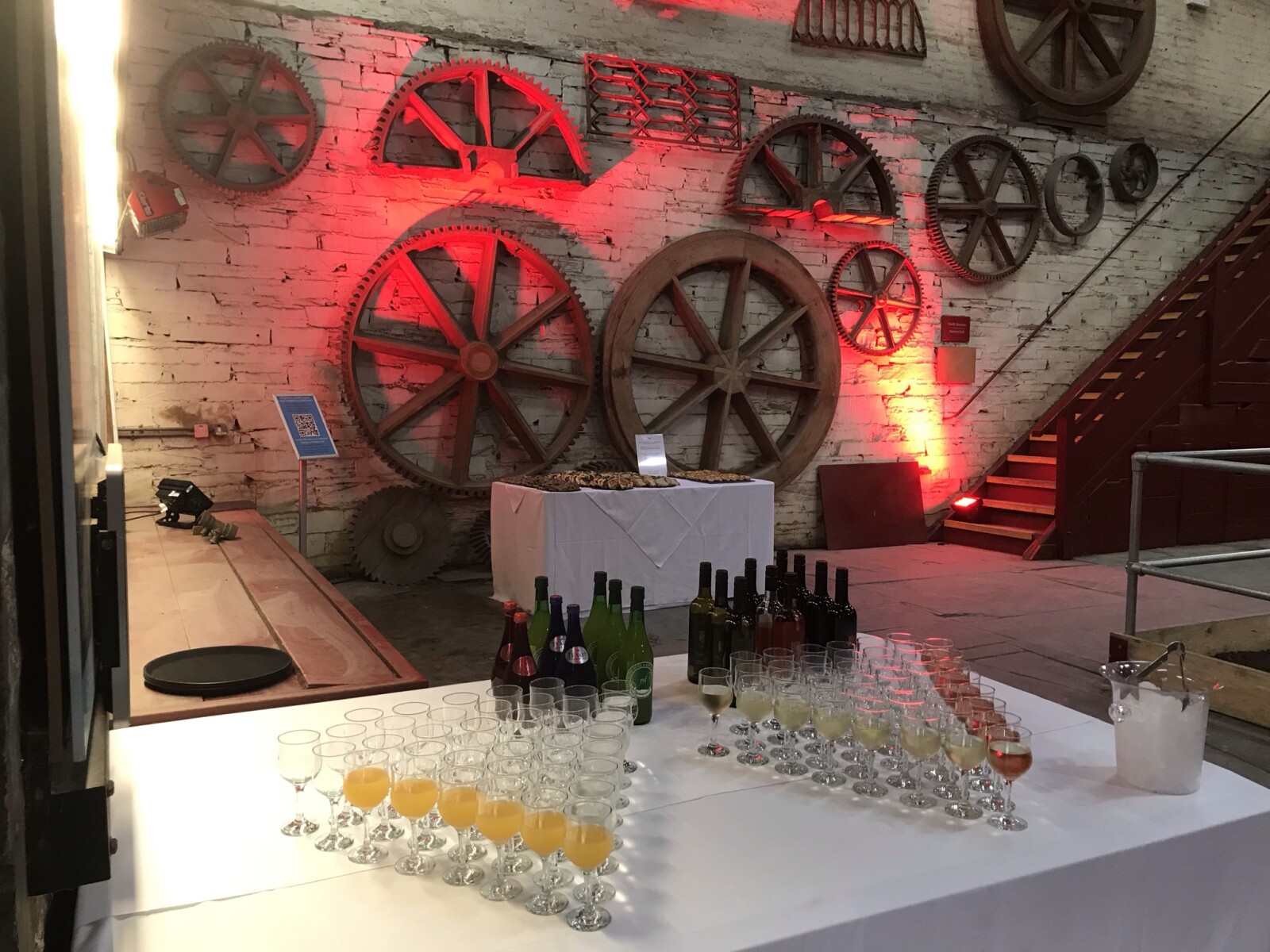 Two triangles of glasses stand on a table with a white tablecloth, some glasses have been filled with wine or orange juice. There's another table of food in the background with a brick wall with different shaped cogs hanging on it.