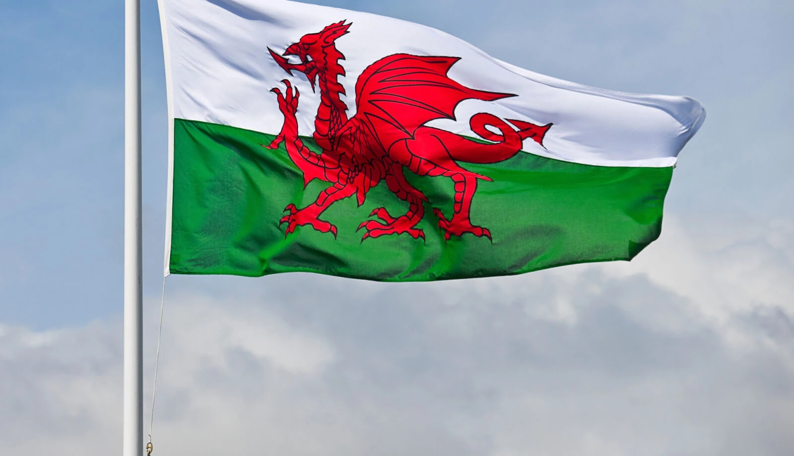 A Welsh flag, featuring a red dragon on a green and white background, flutters in the wind against a cloudy sky.