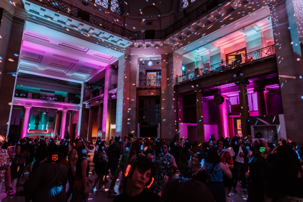 A fairly dark image with the roof of the main hall at NMC lit up with disco lights. There are people dancing whilst wearing multicoloured headphones