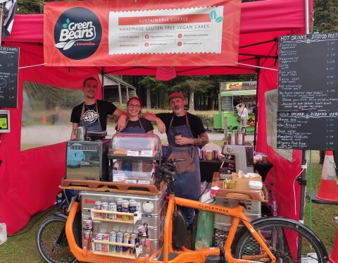 Three people stood behind a coffee counter which is situated in front of an orange bike and there's a red tent around them