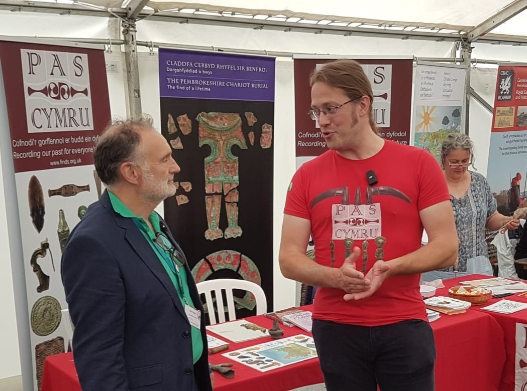 A man in a red tshirt with the words 'Pas Cymru' is stood talking to another man in a blue jacket and green tshirt. They're stood in a marquee and in font of a table with maps on it.