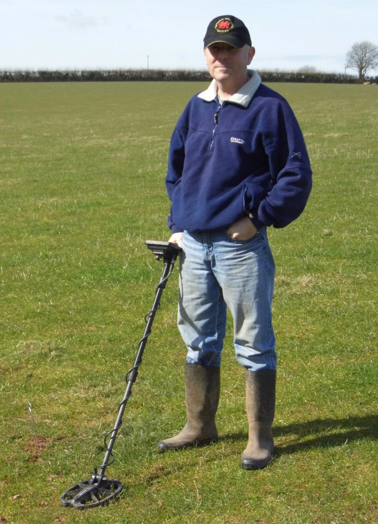  Man stands in a field wearing jeans, navy jumper and a baseball cap, he has one hand in his jeans pocket and the other is holding a metal detector