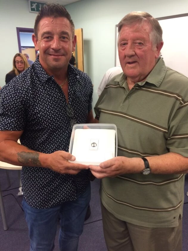 Two men standing next to each other in a classroom, they're both smiling whilst holding an object in a clear container