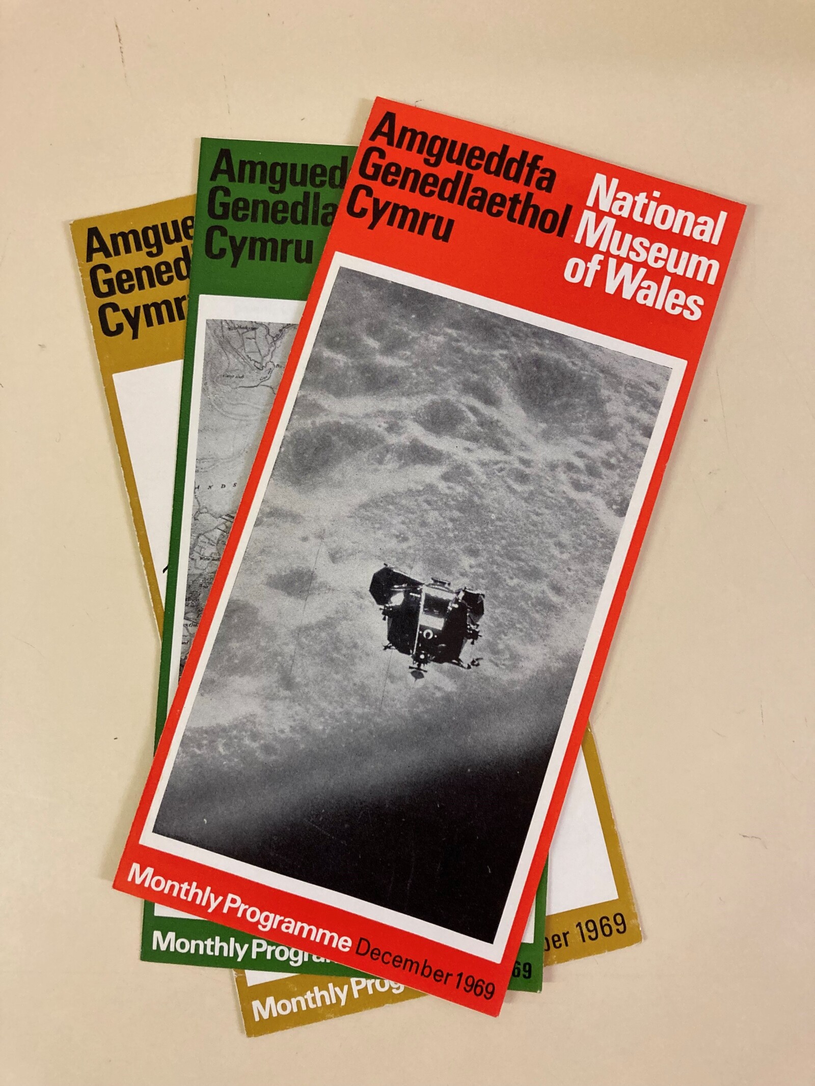 A pile of colourful museum leaflets each with a black and white central photograph. The top leaflet's picture is of the moon and spacecraft.