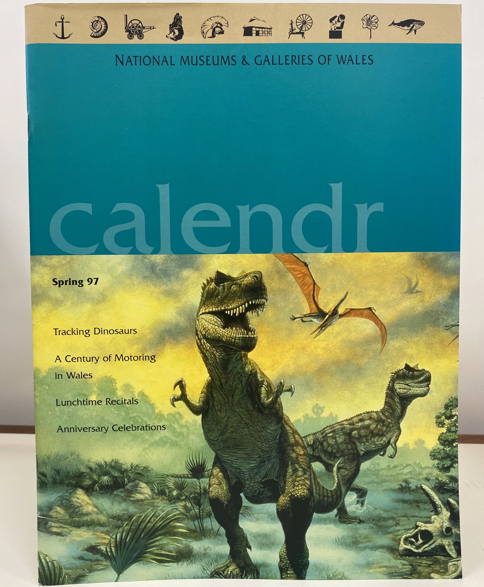 Museum magazine with illustration of dinosaurs. A band of small black icons runs along the top of the magazine 