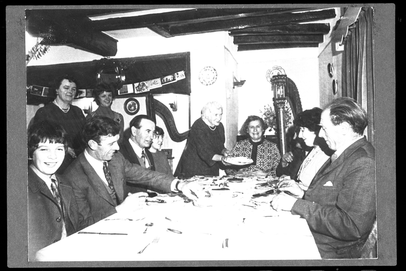 old black and white photograph showing a part of eleven people sat and stood around a large farmehouse table celebrating a Plygain Supper at Cefn Llwyn Farmhouse, Llangynyw