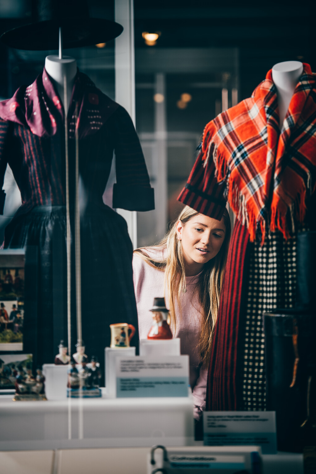 Young woman with blonde hair and a pink jumper looks in awe at items within a collection. Either side there are mannequins with the traditional welsh outfit on them, one with a bright red shawl and the other purple.