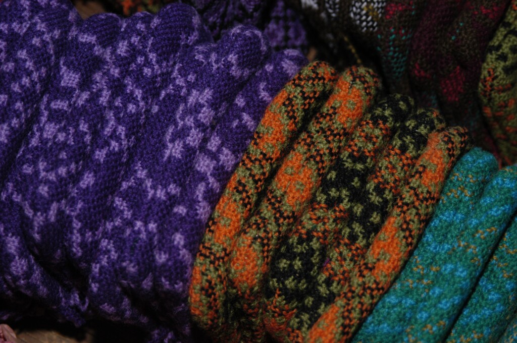 A colourful collection of folded textile materials all packed tightly together. On the left there's purple fabric with a pattern of lighter purple running through it, in the middle there's orange, black and green fabric and on the right green and blue pat