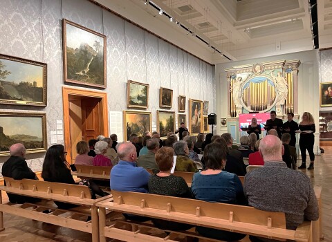 A room full of people sat on benches in front of the organ in a gallery at Amgueddfa Genedlaethol Caerdydd. Four people are stood in front of the audience wearing dark clothing singing