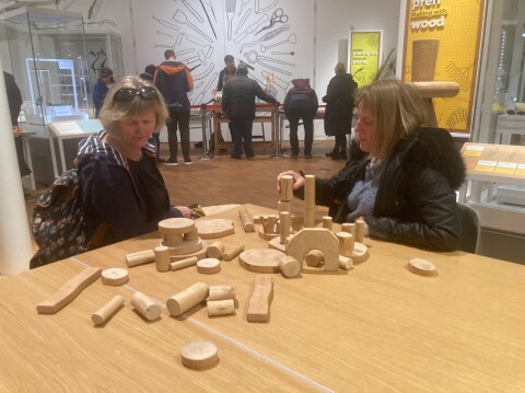 Two women sitting at a table in the Gweithdy Gallery at St Fagans National Museum of History, with some small wooden building blocks in front of them on the table. 