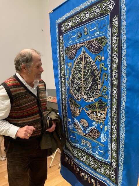 A middle aged man standing in front of a hanging tapestry embroidered in greens and blues, examining it closely. 