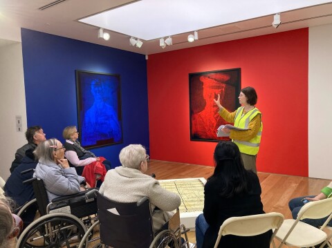A group of adults sitting in a gallery at National Museum Cardiff, some in wheelchairs, listening to a gallery guide tell them about the Artes Mundi 10 exhibition they are visiting. 