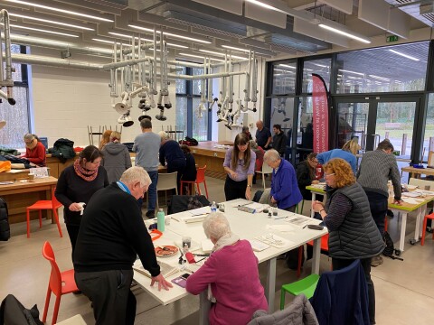 People taking part in the sketching group at St Fagans National Museum of History, sitting and drawing or standing around tables in the workshop space discussing their work.