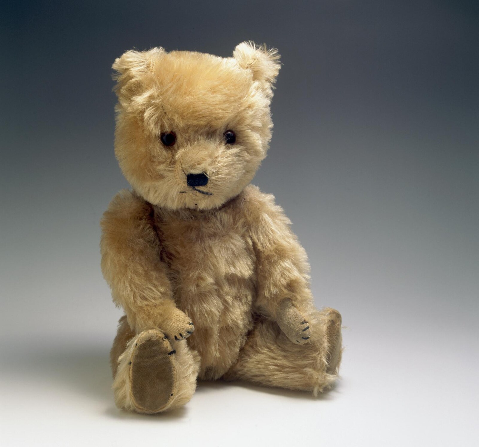 A vintage teddy bear with a light brown, slightly worn fur. The teddy bear is sitting upright against a plain, gradient background that transitions from white at the top to dark grey at the bottom.