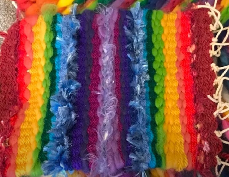 The image shows a colorful, handwoven fabric with rainbow stripes in various textures. The materials range from smooth to fluffy yarns, and loose white strings are visible at the edges.