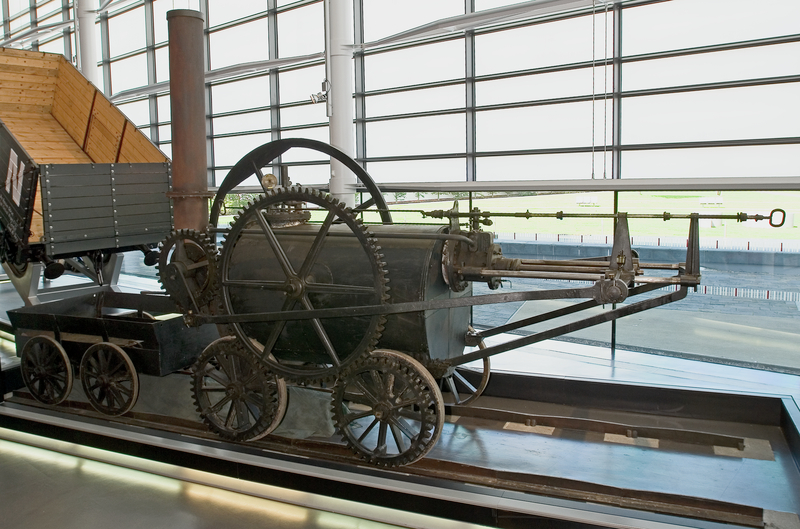 The penydarren locomotive situated at the Nationa Waterfrom Museum. It's sitting on tracks within the museum and there's a tipped trailer behind it 