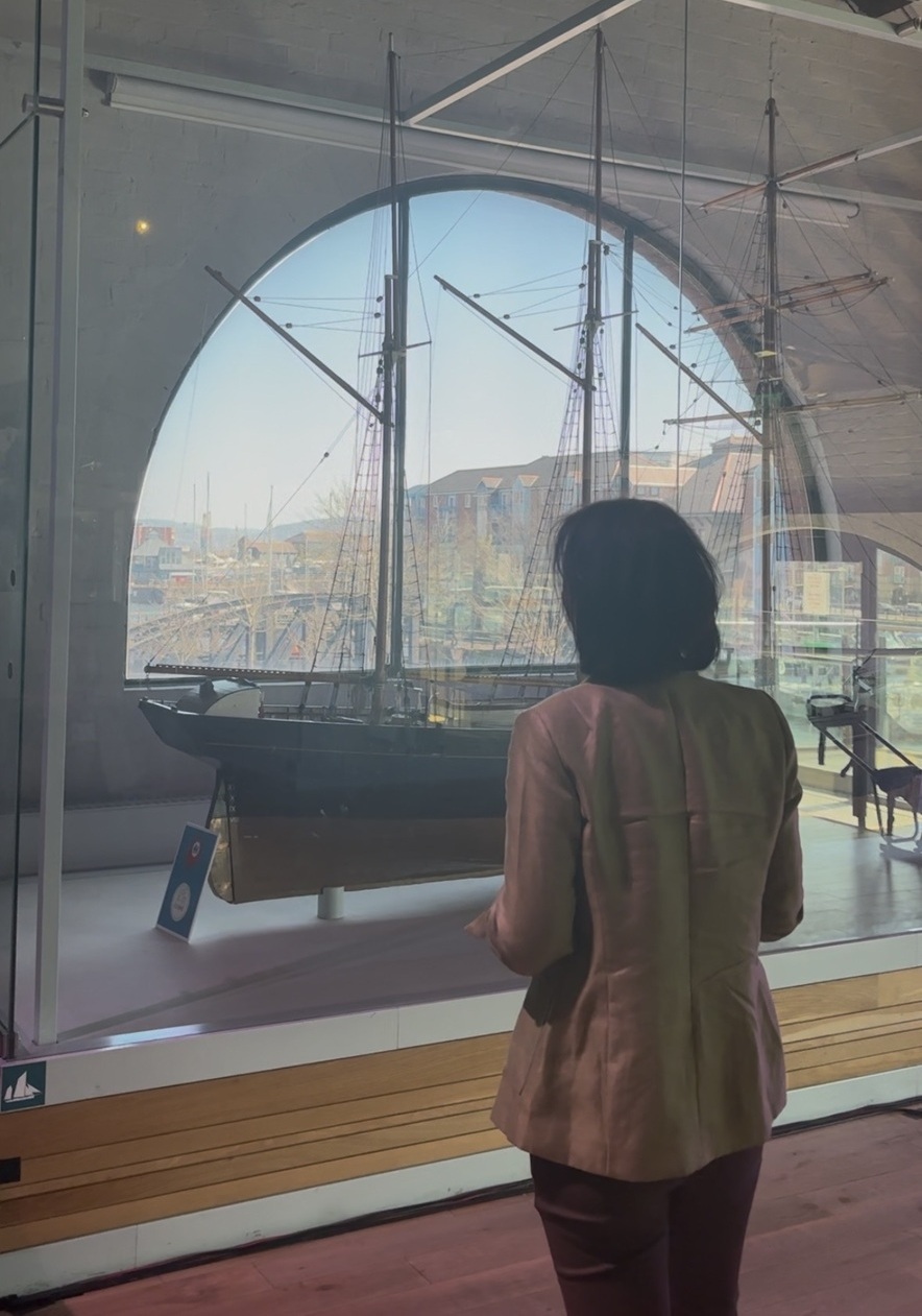 A person stands infront a display cabinet at a museum, within the cabinet there's a ship model. The person has shoulder length hair and is earing a cream leather jacket
