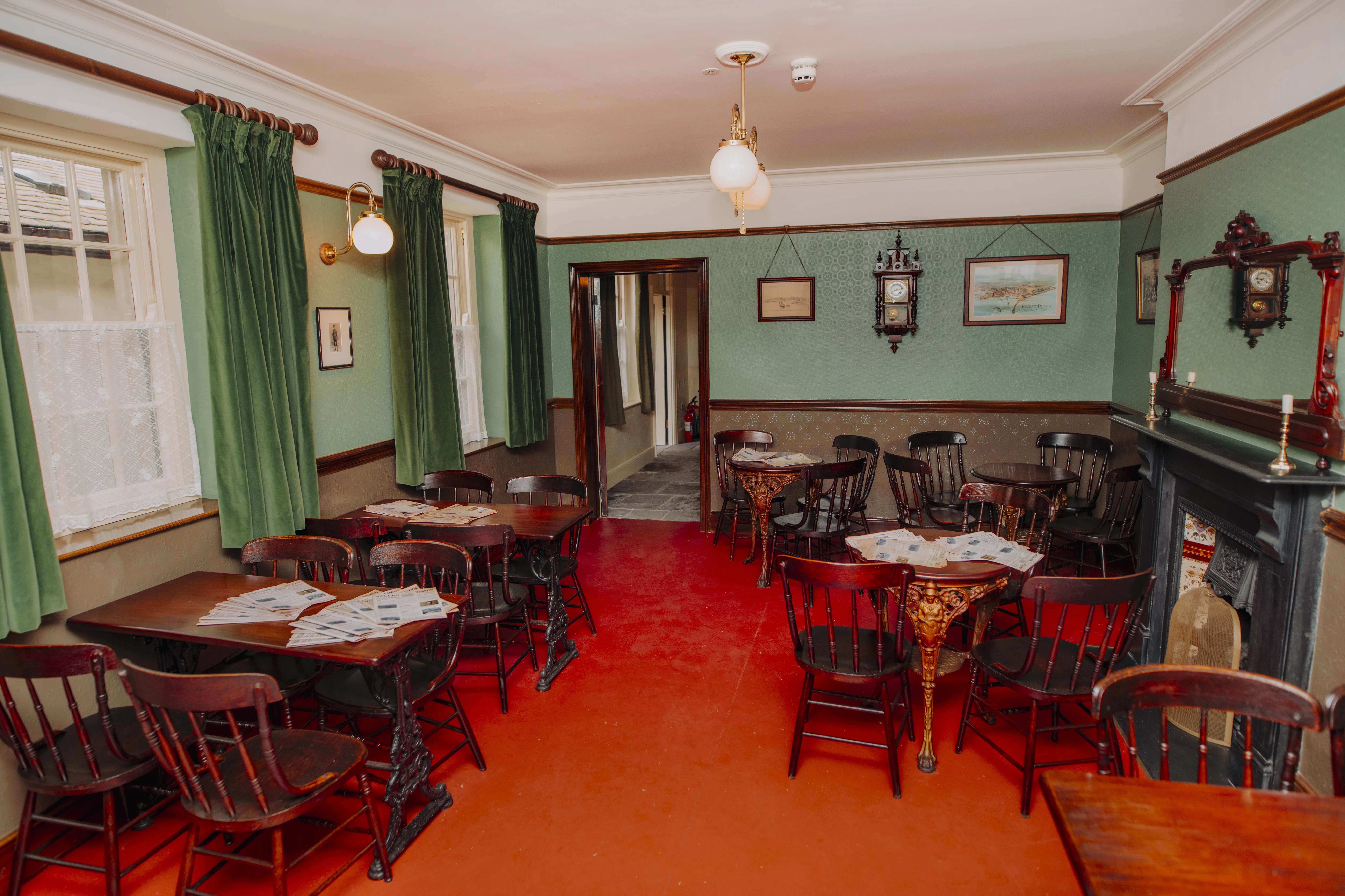 Cosy room with a red floor, green walls and curtains, wooden tables and chairs, a fireplace and framed pictures on the walls.