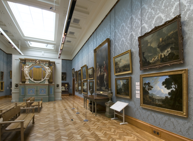 An art gallery with oil paintings hanging on light blue damask walls and an ornate organ case at the far end