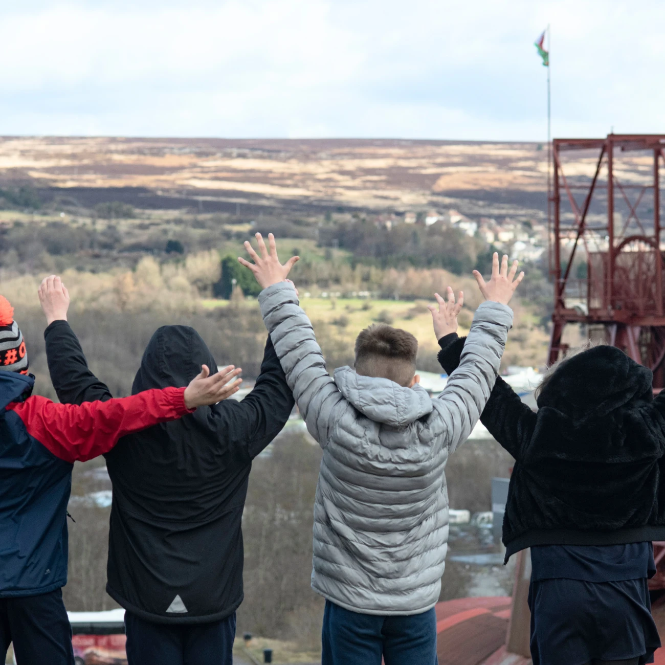 The back of four pupils with their hands in the air. Big Pit head gear can been seen in the distance