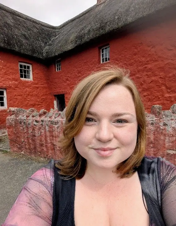 Alt text: A selfie of Kate in front of Kennixton farmhouse, St Fagans. Image shows a women with red hair in a purple dress smiling in front of a red, thatch-roofed, farmhouse. 