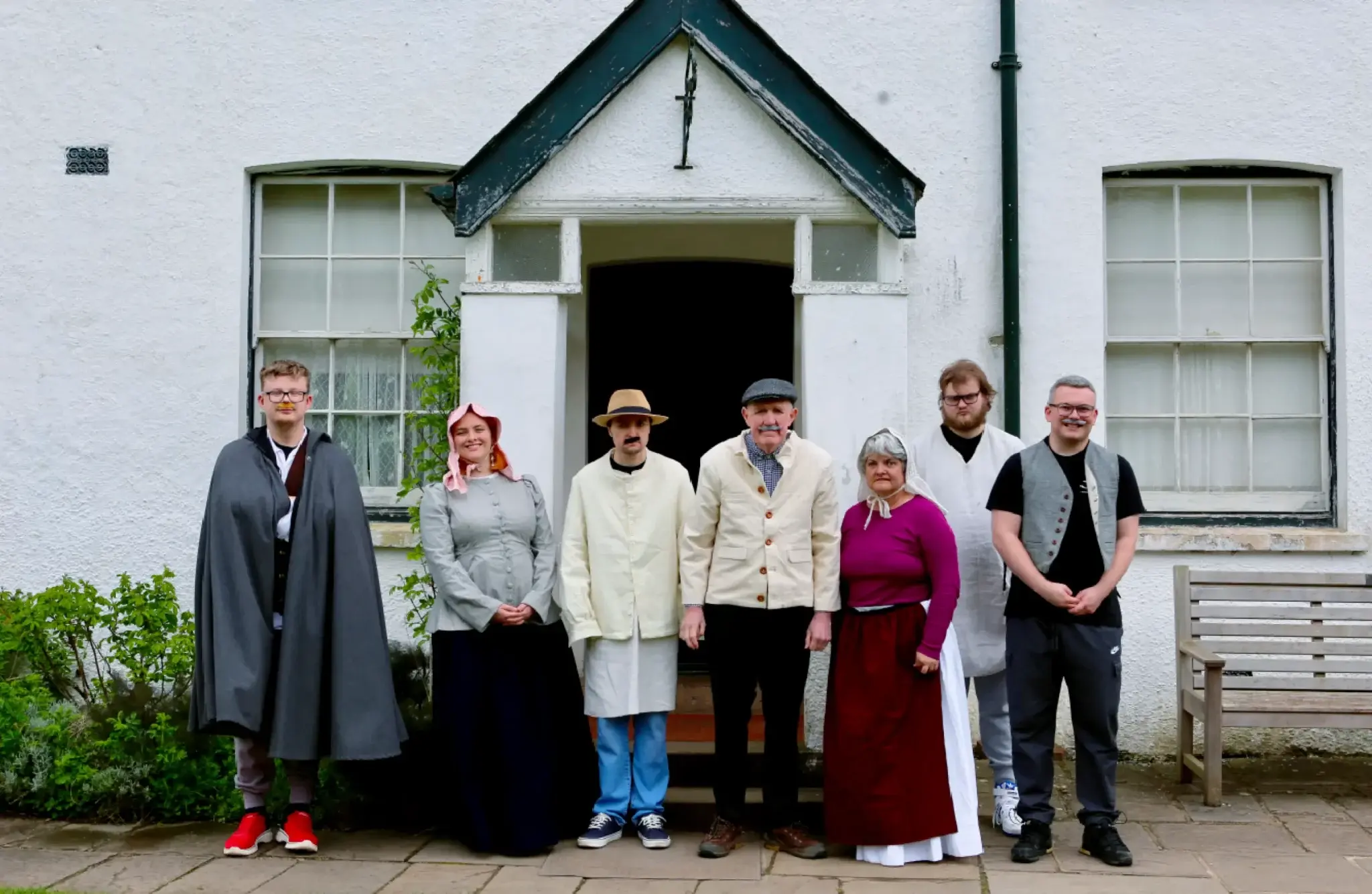 People in a mix of old-fashioned and modern clothes stand together outside a white farmhouse building.